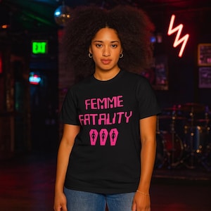 May include: Black t-shirt with the text "FEMME FATALITY" in pink, along with three pink coffin illustrations. The shirt is worn by a person with curly hair, in a dimly lit setting.