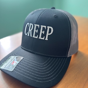 May include: A black trucker hat with a gray mesh back and a curved brim. The word "CREEP" is embroidered in white block letters on the front of the hat. The hat has a Richardson tag.
