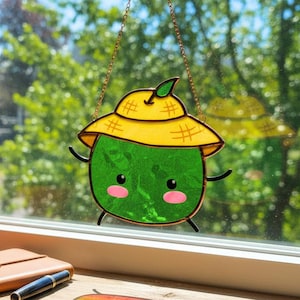 Könnte beinhalten: Ein Glasfenster-Suncatcher mit einer grünen Fruchtfigur mit gelbem Hut. Die Figur hat ein lächelndes Gesicht mit rosa Wangen und hängt an einer goldenen Kette. Der Suncatcher wird vor einem Fenster ausgestellt.