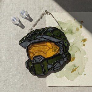 May include: A stained glass helmet in shades of green and yellow, with a black outline and a chain for hanging. The visor is yellow, and the rest of the helmet is green. The design is reminiscent of a video game character.