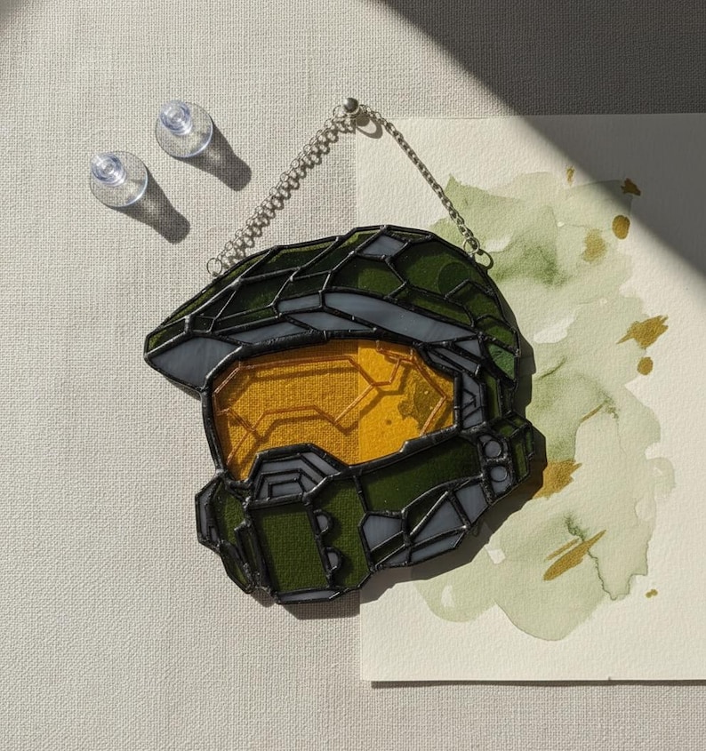 May include: A stained glass helmet in shades of green and yellow, with a black outline and a chain for hanging. The visor is yellow, and the rest of the helmet is green. The design is reminiscent of a video game character.