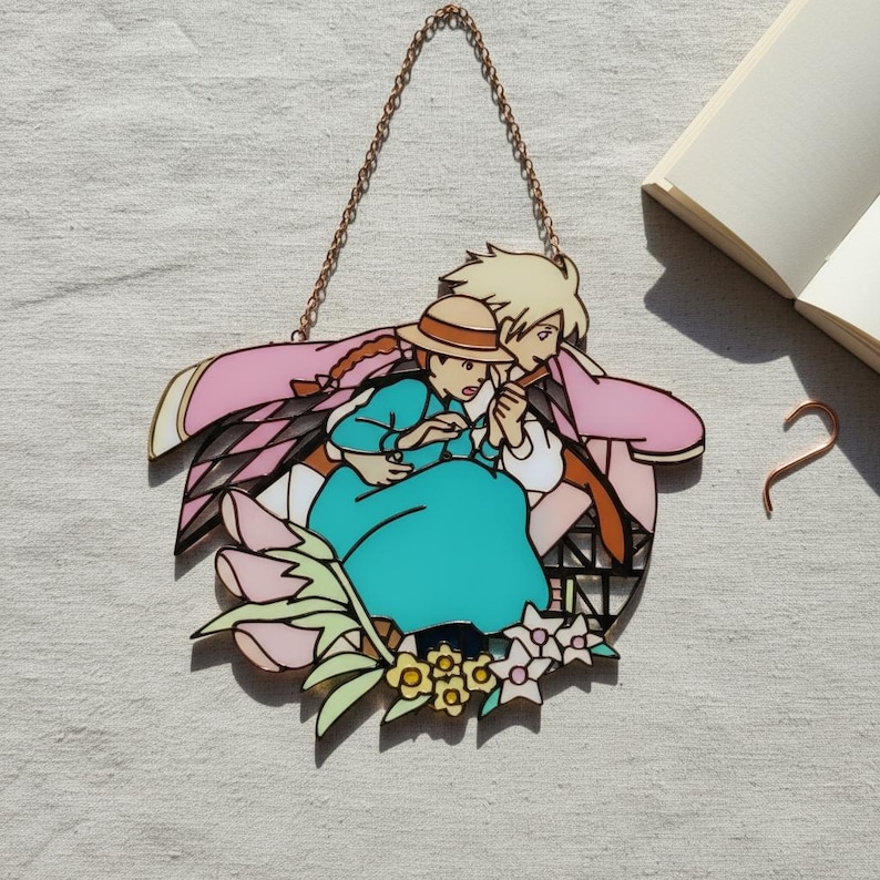 May include: A stained glass panel featuring a couple from the animated film, Howl's Moving Castle. The artwork depicts a man and woman in a loving embrace, surrounded by pink and black accents, with a copper chain for hanging.