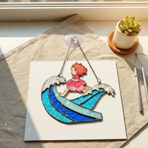 May include: Stained glass art piece depicting a girl riding a wave. The wave is composed of shades of blue and white glass. The girl is wearing a pink dress and has red hair. The piece is suspended by a chain.