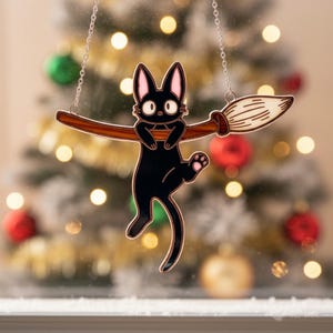 May include: A black cat ornament with pink ears and a white face, hanging from a silver chain. The cat is holding onto a brown broom with a white brush. The background is blurred with Christmas tree lights and ornaments.
