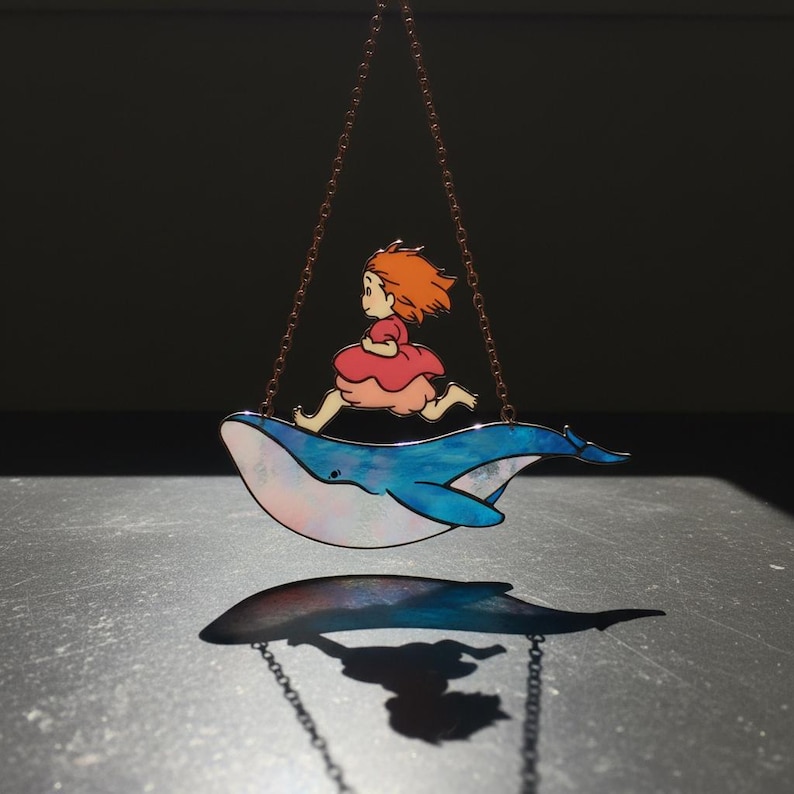 May include: A whimsical hanging decoration featuring a girl with orange hair running on a blue whale. The whale has iridescent accents. The girl wears a pink dress. The decoration is suspended by a chain. The shadow of the whale is visible below.