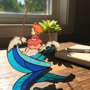 May include: A stained glass suncatcher featuring a cartoon character with orange hair and a red dress running on a blue and white wave. The suncatcher is suspended by a copper chain. A succulent plant and a notebook are in the background.