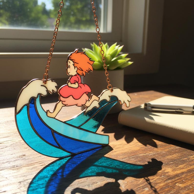 May include: A stained glass suncatcher featuring a cartoon character with orange hair and a red dress running on a blue and white wave. The suncatcher is suspended by a copper chain. A succulent plant and a notebook are in the background.
