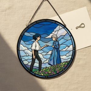 May include: Round stained glass art piece featuring a scene with two figures in a blue sky and cloud background. The figures are in a field of flowers. The artwork is framed in black and hangs from a chain.