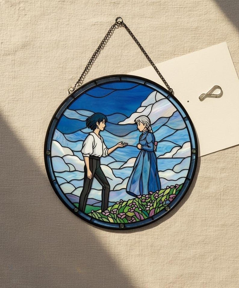May include: Round stained glass art piece featuring a scene with two figures in a blue sky and cloud background. The figures are in a field of flowers. The artwork is framed in black and hangs from a chain.