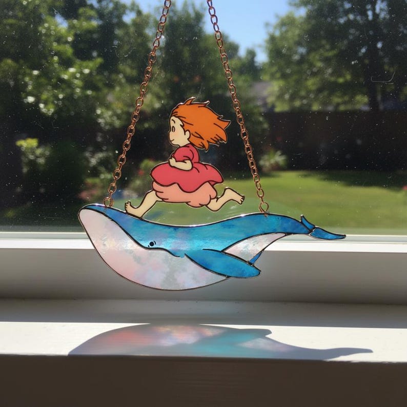 May include: A stained glass window hanging featuring a cartoon girl in a pink dress running on the back of a blue whale. The whale is suspended by a copper chain, with a sunny outdoor background.