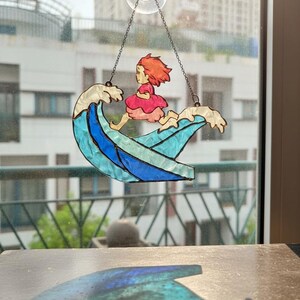 May include: Stained glass suncatcher depicting a girl in a pink dress riding a wave. The wave is blue and white, and the girl has red hair. The suncatcher hangs from a chain and suction cup.