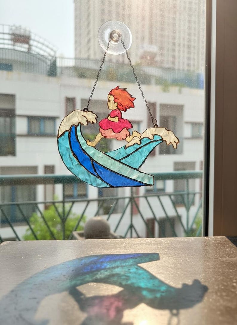 May include: Stained glass suncatcher depicting a girl in a pink dress riding a wave. The wave is blue and white, and the girl has red hair. The suncatcher hangs from a chain and suction cup.