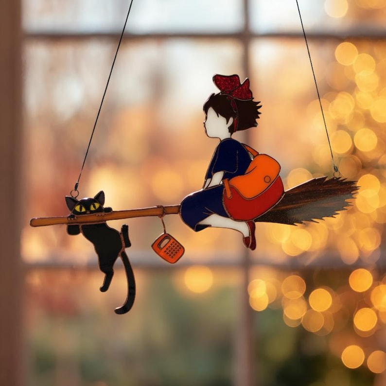 May include: A whimsical stained glass suncatcher featuring a young witch riding a broom with a black cat. The witch wears a blue dress, red bow, and carries an orange backpack. A small orange radio hangs from the broom. The suncatcher is suspended by a black string.