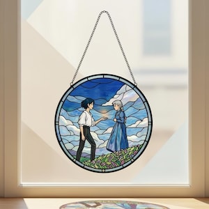 May include: Round stained glass window art featuring two figures in a landscape with a blue sky and clouds. The artwork is framed in black and hangs from a chain. The figures are in a field of flowers.