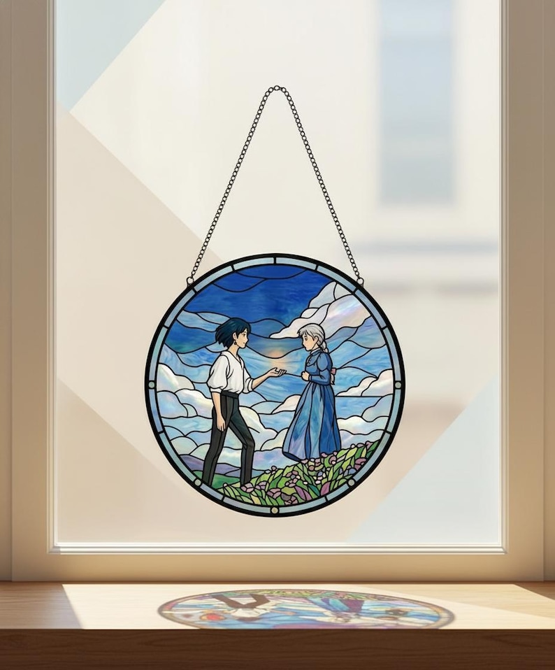 May include: Round stained glass window art featuring two figures in a landscape with a blue sky and clouds. The artwork is framed in black and hangs from a chain. The figures are in a field of flowers.
