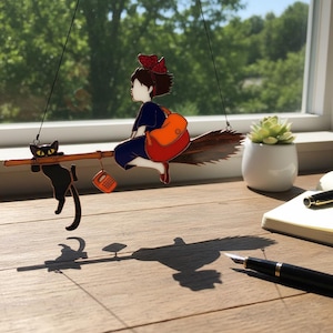 May include: A whimsical hanging decoration featuring a young witch riding a broom with a black cat. The witch wears a blue dress, red shoes, and a red bow. An orange bag hangs from the broom. The cat hangs from the broom. A small orange phone hangs from the broom.