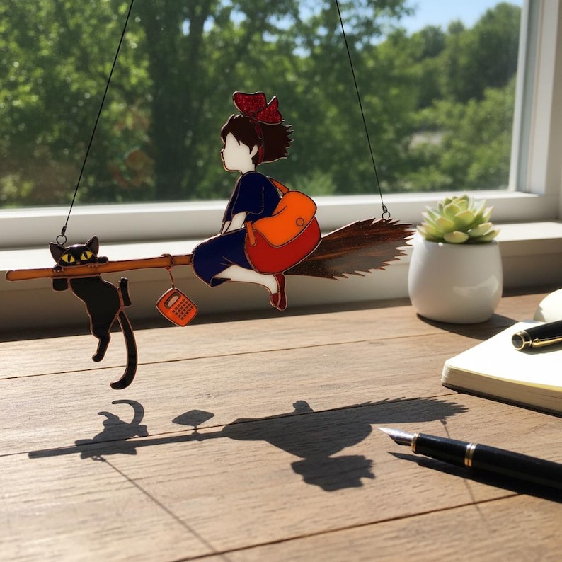 May include: A whimsical hanging decoration featuring a young witch riding a broom with a black cat. The witch wears a blue dress, red shoes, and a red bow. An orange bag hangs from the broom. The cat hangs from the broom. A small orange phone hangs from the broom.