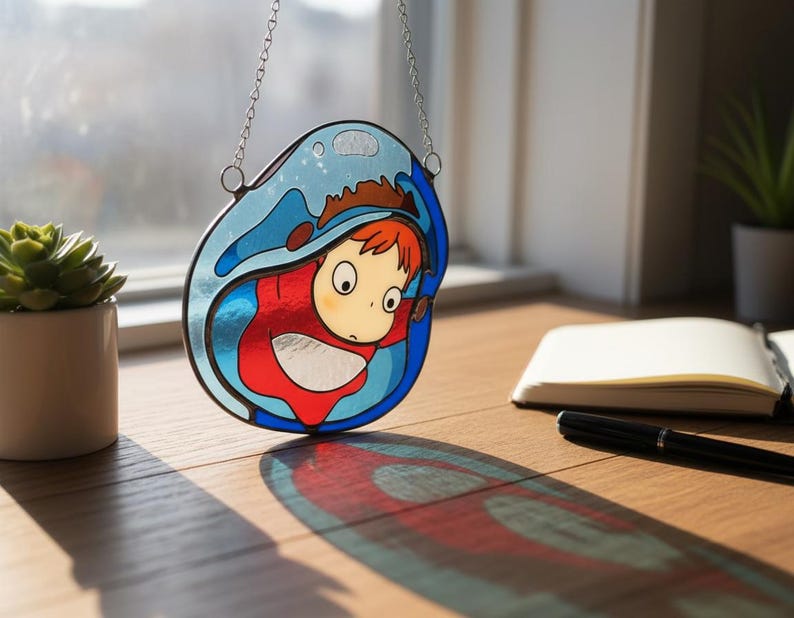 May include: Stained glass art piece featuring a cartoon character in a red outfit, surrounded by blue water. The artwork is suspended by a silver chain. The piece casts a colorful shadow on a wooden surface, with a succulent plant and notebook in the background.