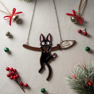 May include: A black cat pendant necklace with a brown broom design. The cat has pink ears and paws. The necklace is on a silver chain. The background includes Christmas bells, red berries, and evergreen branches.