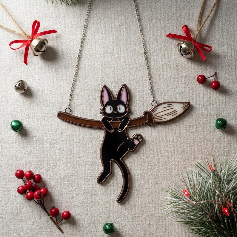 May include: A black cat pendant necklace with a brown broom design. The cat has pink ears and paws. The necklace is on a silver chain. The background includes Christmas bells, red berries, and evergreen branches.