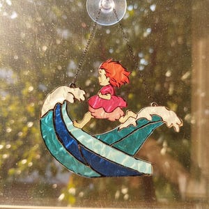 May include: Stained glass window art featuring a girl with red hair in a pink dress riding a wave. The wave is blue and teal, with white foam. The artwork is suspended by a chain and suction cup.