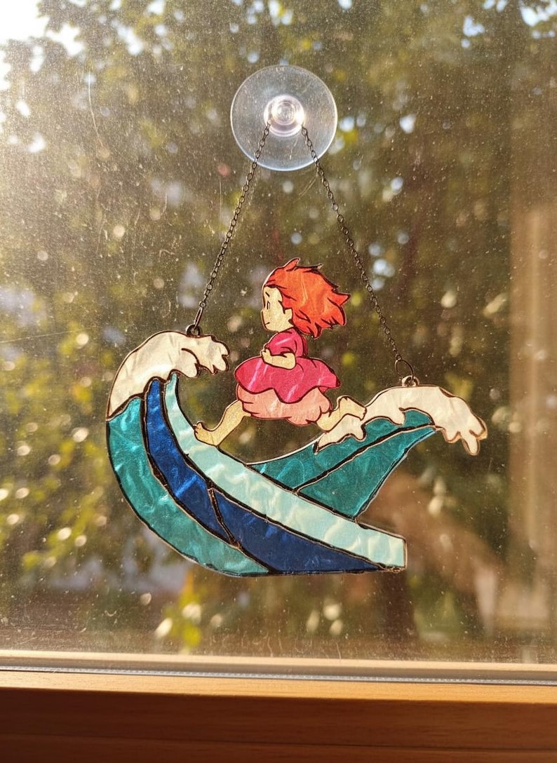May include: Stained glass window art featuring a girl with red hair in a pink dress riding a wave. The wave is blue and teal, with white foam. The artwork is suspended by a chain and suction cup.