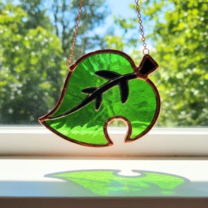 May include: A stained glass leaf in shades of green, with a black accent and copper-colored trim. The leaf is suspended by a delicate copper chain. The design casts a shadow on the white surface below.