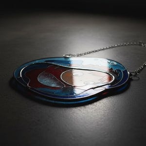 May include: Oval-shaped pendant with a silver chain. The pendant features a design with blue, red, and white colors. The artwork appears to depict a person or figure. The pendant is likely made of metal and glass.