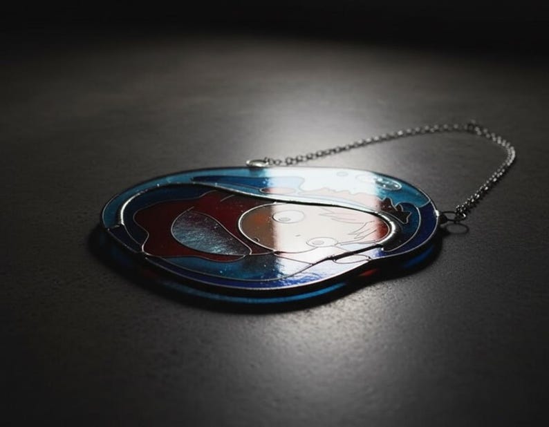 May include: Oval-shaped pendant with a silver chain. The pendant features a design with blue, red, and white colors. The artwork appears to depict a person or figure. The pendant is likely made of metal and glass.