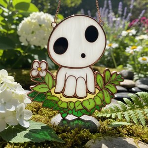以下が含まれることがあります： 大きな黒い目と小さな口を持つ、白い精霊のような姿を描いたステンドグラスのサンキャッチャー。緑の葉と小さな花に囲まれ、銅色の縁と吊り下げチェーンが付いています。サンキャッチャーは屋外に展示されています。