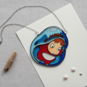 May include: A silver chain necklace with a pendant featuring a cartoon character in a red outfit, framed by blue and clear glass, resembling a wave. The pendant is on a white surface with a piece of driftwood and small white shells.