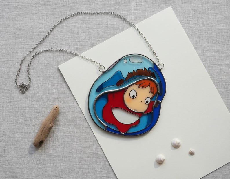 May include: A silver chain necklace with a pendant featuring a cartoon character in a red outfit, framed by blue and clear glass, resembling a wave. The pendant is on a white surface with a piece of driftwood and small white shells.