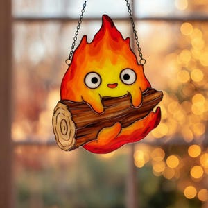 May include: A stained glass decoration depicting a cartoon fire spirit holding a log. The spirit is in shades of yellow, orange, and red, with black eyes and a smiling face. It hangs from a chain, suitable for a window or wall.