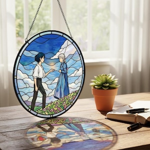 May include: Round stained glass window art depicting two figures in a landscape with a blue sky and clouds. The artwork is framed in black and hangs from a chain. A small potted succulent and a notebook are on a wooden table.