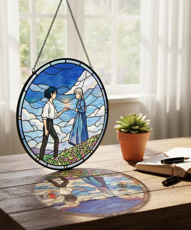 May include: Round stained glass window art depicting two figures in a landscape with a blue sky and clouds. The artwork is framed in black and hangs from a chain. A small potted succulent and a notebook are on a wooden table.