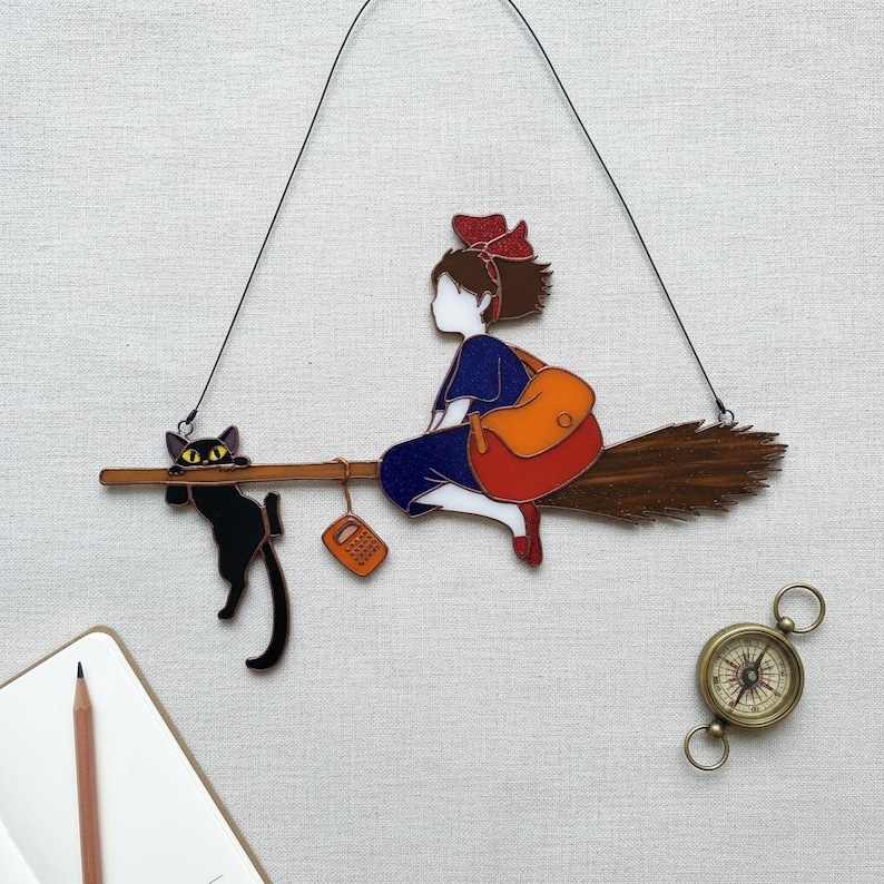 May include: A whimsical wall hanging featuring a girl in a blue dress riding a broom with a black cat. The girl has a red bow and an orange backpack. A small orange phone hangs from the broom. A compass is at the bottom right.