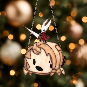 Może przedstawiać: Szklana ozdoba z postaciami z gry wideo Hollow Knight. Ozdoba przedstawia Hornet stojącą na stylizowanej postaci. Zawieszona na łańcuszku, na rozmytym tle światełek choinkowych, co tworzy świąteczną atmosferę.