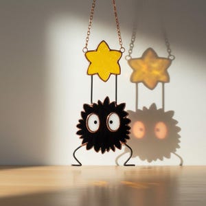 May include: A whimsical stained glass suncatcher featuring a yellow star above a black, spiky creature with large white eyes. The suncatcher hangs from a copper chain and casts a shadow on the wall. The design is reminiscent of a cartoon character.