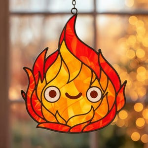 May include: A stained glass suncatcher shaped like a cartoon flame with a smiling face. The flame is made of orange and red glass, outlined in black. It hangs from a small chain.