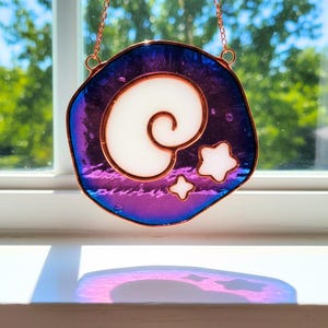 Può includere: Un acchiappasole in vetro colorato con un bordo color rame. Il design presenta una spirale bianca e due stelle bianche su uno sfondo iridescente viola e blu. L'acchiappasole è appeso a una catena di rame.