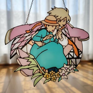 May include: Stained glass art piece depicting two figures from an animated film, with a teal dress, pink cape, and a straw hat. The artwork is accented with floral elements and a chain for hanging. The piece is a decorative item.