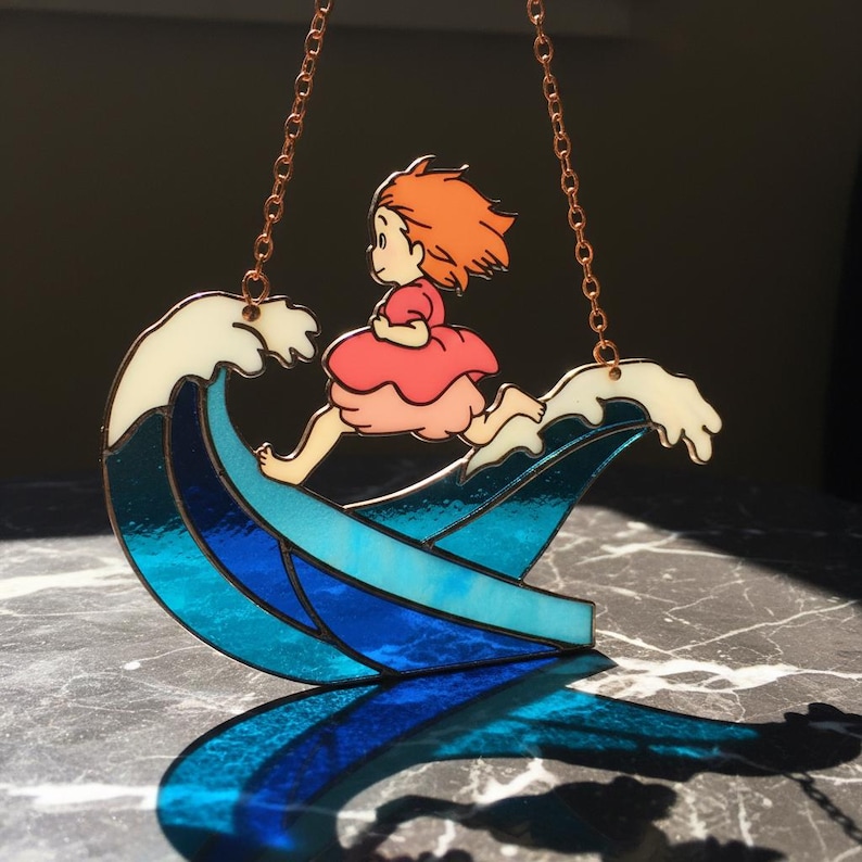 May include: A stained glass suncatcher featuring a cartoon girl with orange hair and a pink dress running on a blue and white wave. The suncatcher is suspended by copper-colored chains.