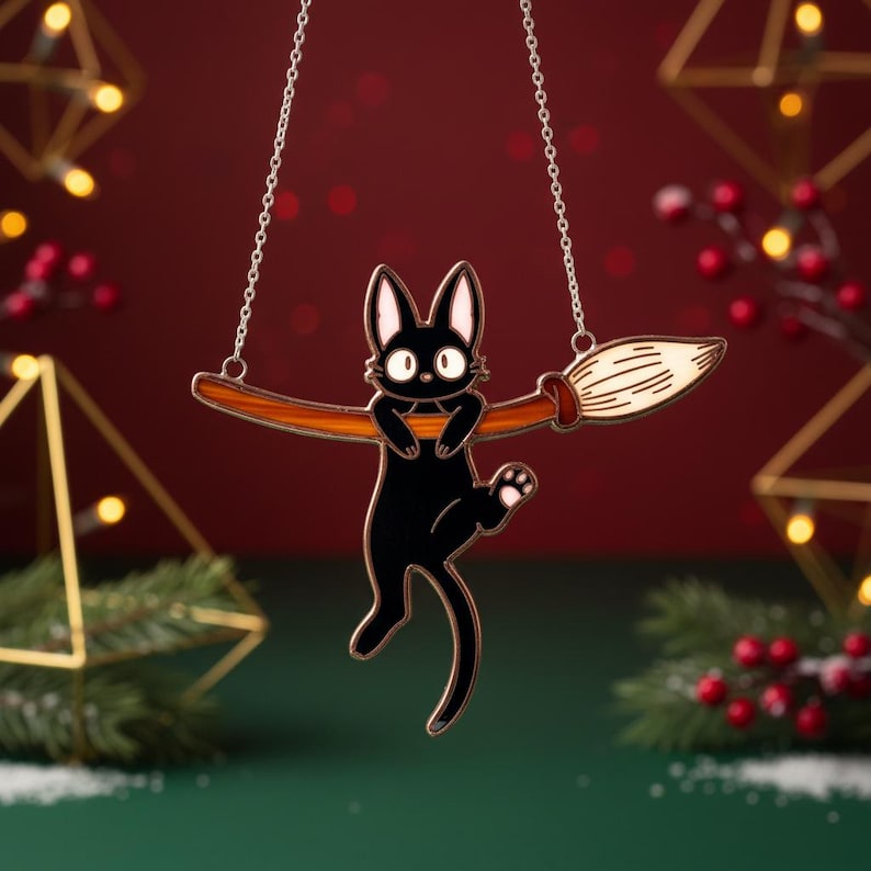 May include: A black cat pendant with a white face and paws, riding a brown broom. The cat is suspended from a silver chain. The background is a deep red with gold geometric shapes and festive greenery.