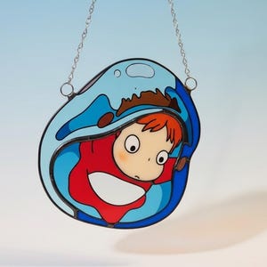May include: Stained glass suncatcher featuring a cartoon character in a red outfit, surrounded by blue water. The artwork is outlined in black and hangs from a silver chain. The character has large eyes and orange hair.