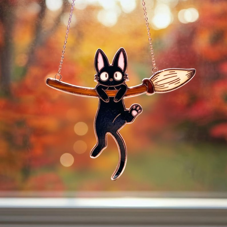 May include: A black cat-shaped suncatcher hanging from a chain, perched on a brown broom. The cat has large, round eyes and is suspended against a blurred background of fall colors. The broom's bristles are detailed.