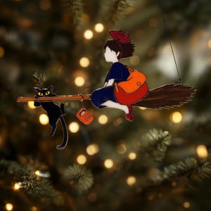 May include: A whimsical ornament featuring a young witch riding a broom with a black cat and an orange bag. The witch is wearing a blue dress and a red bow. The cat is hanging on the broom. The ornament is suspended by a thin wire.
