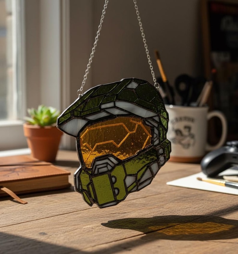 May include: A stained glass Spartan helmet in green and gold, from the Halo video game series, suspended by a silver chain. The helmet has a yellow visor. The background is blurred, showing a window and desk.