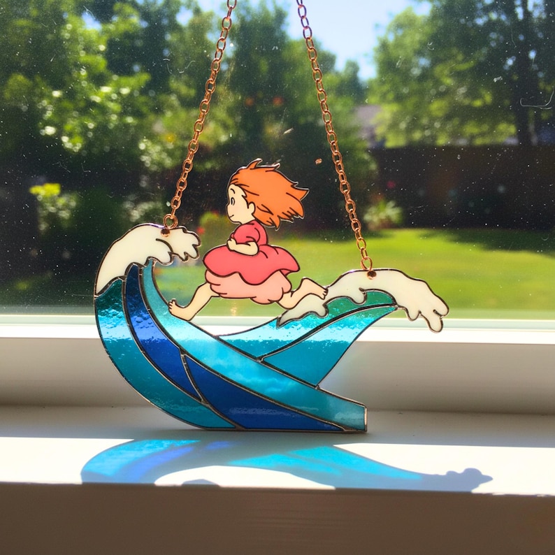 May include: A stained glass suncatcher featuring a cartoon girl running on a wave. The wave is blue and white, with a copper chain for hanging. The girl has red hair and is wearing a pink dress. The suncatcher is displayed in front of a window.