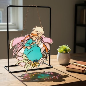May include: A stained glass panel featuring a cartoon couple in a loving embrace, with a pink and white patterned background. The panel is suspended from a black metal frame by a gold chain. The couple is wearing a blue dress and a hat.