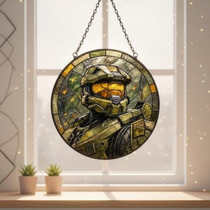 May include: A circular stained glass panel depicting a character in green armour. The artwork is suspended by a chain and set against a window. The character's visor glows with a warm, golden light, set against a backdrop of green.